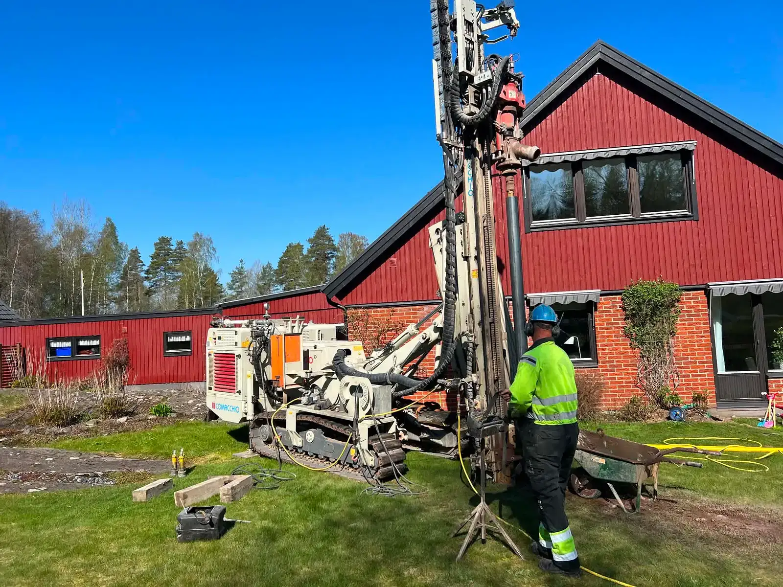 En arbetare med borrigg utför geotermisk borrning vid ett rött hus.